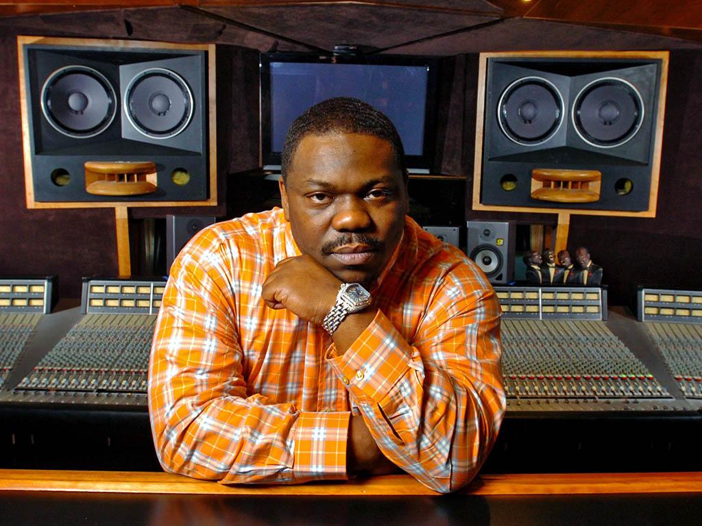 A man in an orange checkered shirt sitting in front of a large music mixing console with studio monitors in the background.