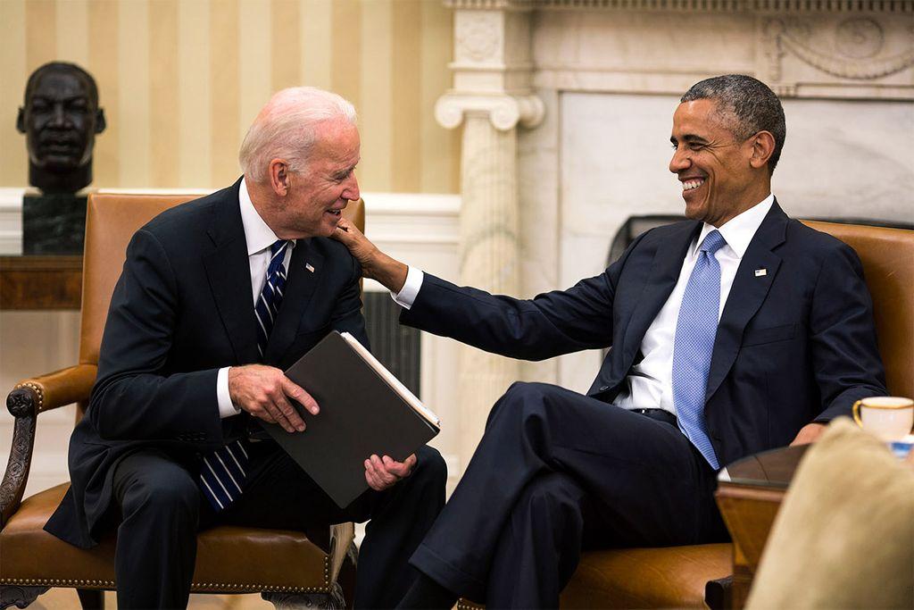 Former President Barack Obama and current President Joe Biden share a light moment in the Oval Office, with Obama smiling while lightly touching Biden's shoulder.
