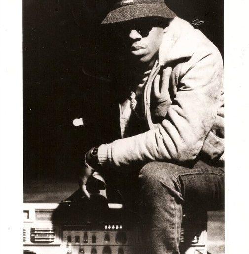A black and white image of a man wearing sunglasses and a bucket hat, sitting in front of a sound system. He is dressed in a shearling-lined jacket and jeans.