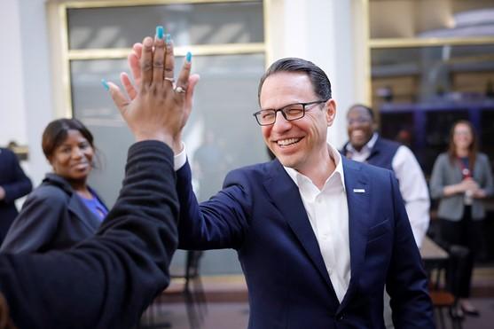 A man in a suit smiling and giving a high five to another person, surrounded by smiling individuals in an indoor setting.