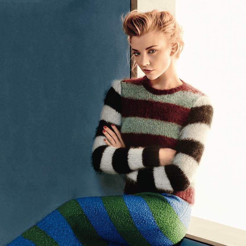A woman poses against a backdrop with a blue wall, wearing a colorful striped sweater and a matching patterned skirt, showcasing a stylish and cozy fashion look.