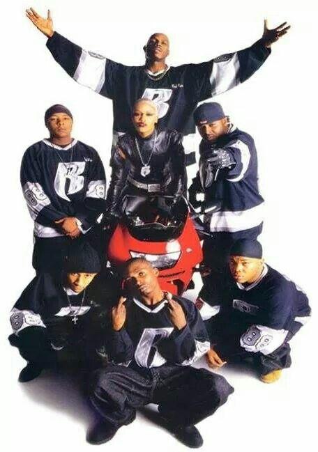 A group of seven musicians posing together, wearing matching black and white outfits, in front of a red motorcycle. The central figure is standing with arms raised while others are crouched or kneeling around.
