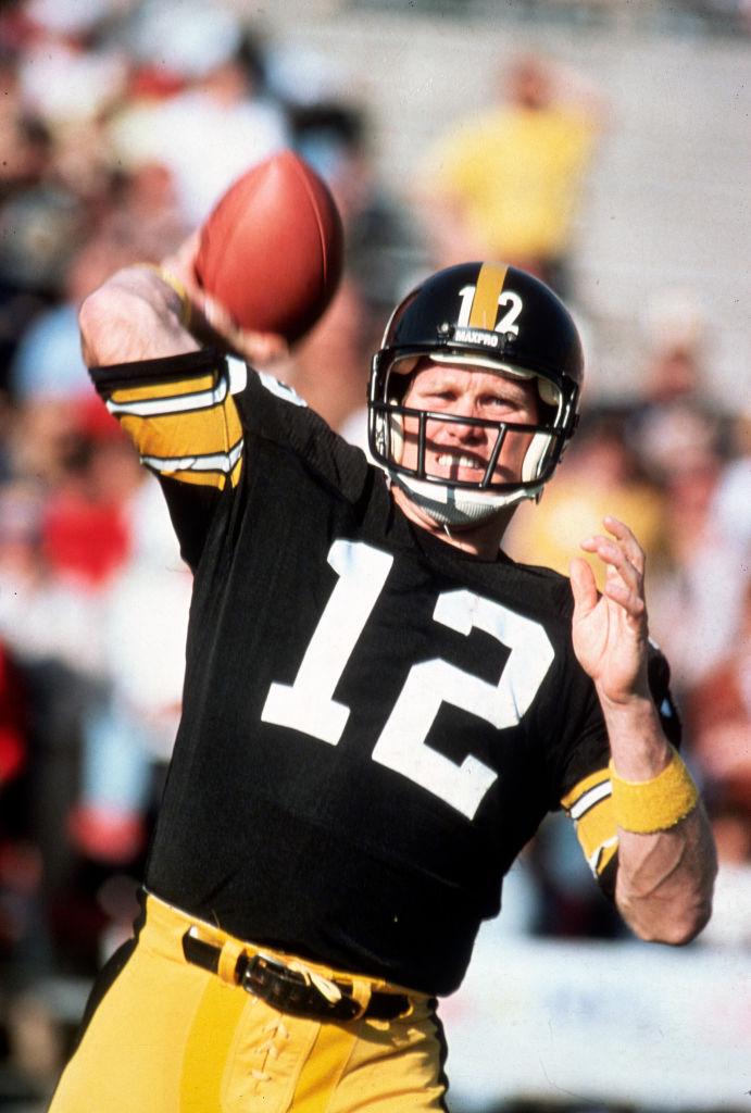 A football player in a black jersey with the number 12 and yellow pants throwing a football during a game.