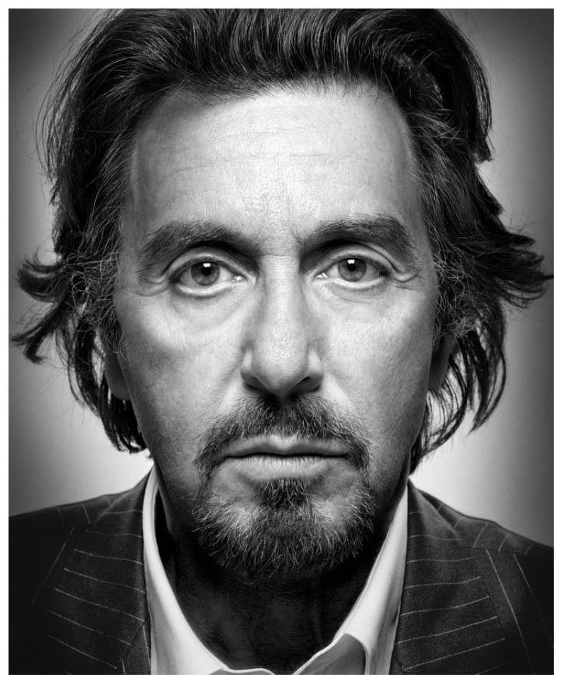 Close-up portrait of a man with long dark hair and a beard, wearing a pinstripe suit and white shirt, set against a grey background.