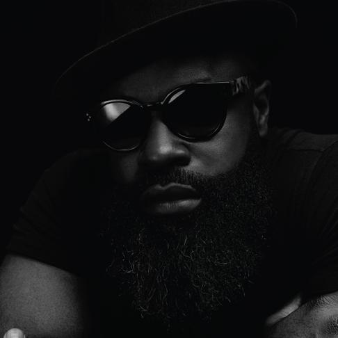 A man with a thick beard, wearing dark sunglasses and a black hat, poses against a black background.