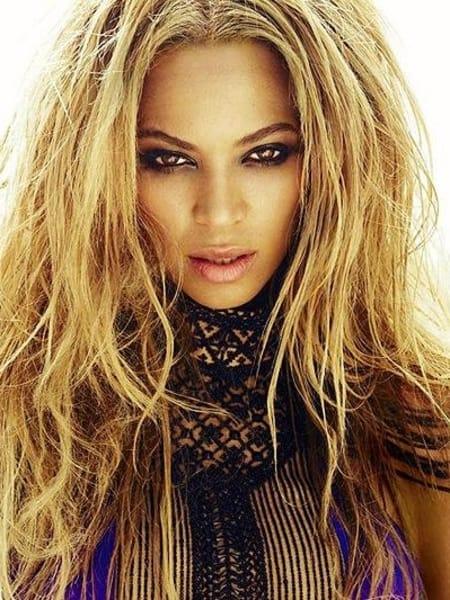 Close-up portrait of a person with long, wavy, blonde hair and striking makeup, looking directly at the camera.