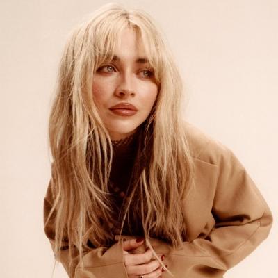 A woman with long blonde hair, wearing a beige oversized jacket, poses with a thoughtful expression against a soft, neutral background.