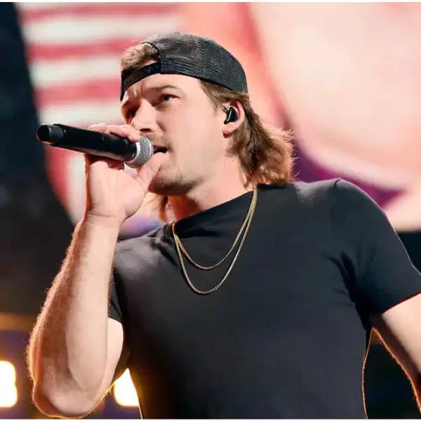 A male singer with long hair wearing a black cap and t-shirt, holding a microphone while performing on stage.